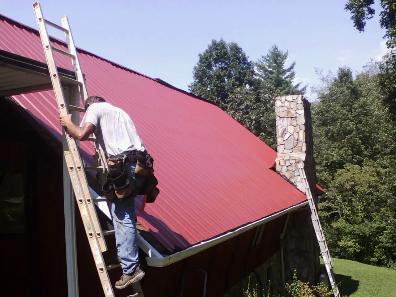 Roofer on a ladder beside a red metal roof and stone chimney — Soffit & Fascia Repair by Roofing Brothers in Versailles